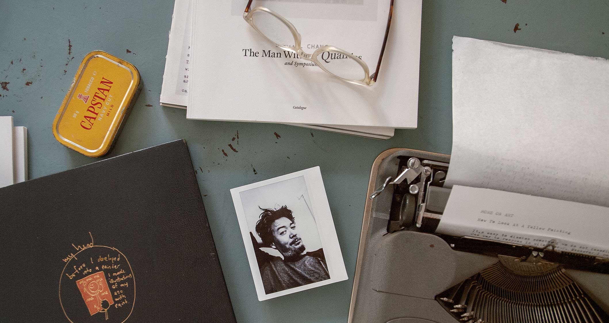Tabletop with a polaroid of the artist, catalogues, glasses, typewriter and a sheet of paper entitled More On Art